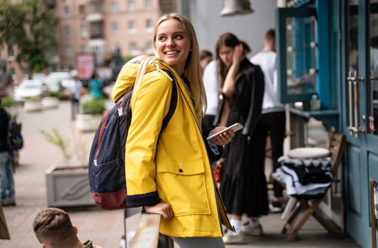 Jente i gul regnjakke lener seg mot et gjerde på fortauet. Hun smiler og holder en telefon i hånden.